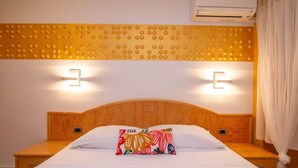 Habitación de hotel con cabecera de madera, dos mesitas de noche, cama con ropa blanca, dos lámparas de pared y un equipo de aire acondicionado.