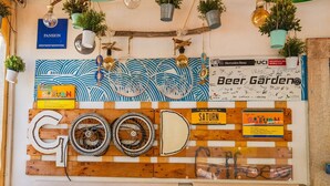 Una pared con diversos letreros y adornos, incluyendo un gran letrero que dice 'GOOD' hecho con tablones de madera y ruedas de bicicleta, rodeado de plantas en macetas y bombillas colgantes.
