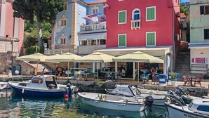 A marina with boats docked, a red building with green shutters, and outdoor seating with umbrellas. - Pansion Saturn / Pansion Saturn soba 16 - seaview (Veli Lošinj)