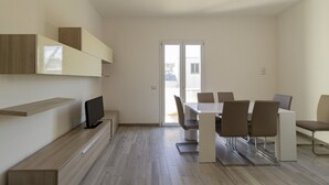 Un comedor moderno y minimalista con una mesa blanca y sillas marrones, un televisor en un mueble bajo y un amplio ventanal corredizo que da a un balcón.