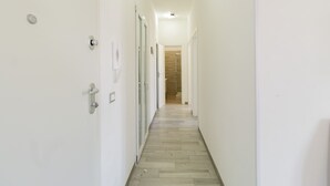Un pasillo estrecho con paredes blancas y suelo de madera, que conduce a una habitación con puerta de madera e interruptor de luz en la pared.
