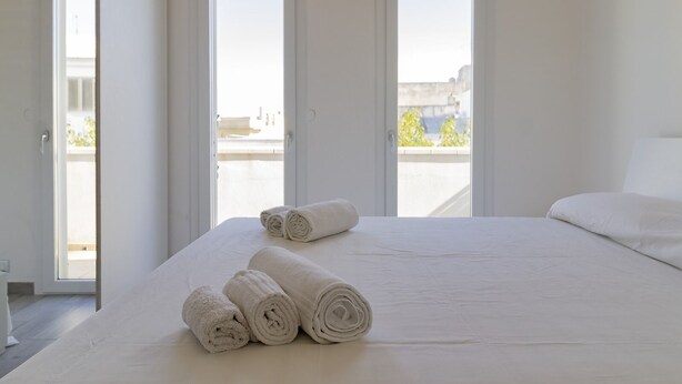 Una habitación de hotel con una cama grande, tres toallas enrolladas y tres ventanas con vistas a la ciudad.