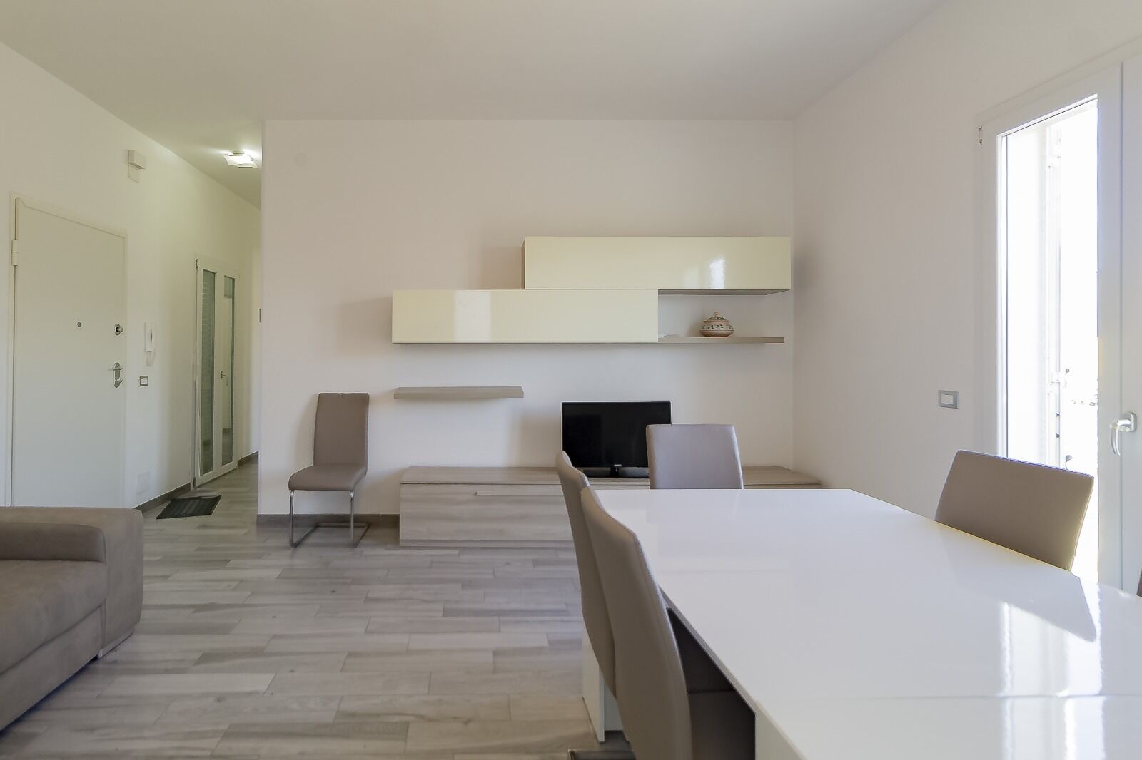 A modern, minimalist living room with a large white table, two chairs, a flat-screen TV, and built-in shelving.