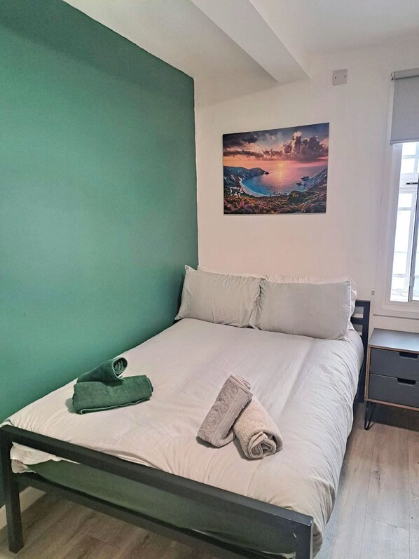 A neatly made bed with a green towel and a rolled-up towel, a painting of a sunset on the wall, and a window with blinds. - Practical & Affordable 2 BR Flat in City Centre (Sheffield)