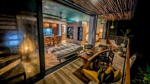 A modern interior with a sofa, dining table, and chairs. There is a kitchen area with a stove and cabinets. The room has a wooden floor and a large window. - Nuju Rentals|1Br Penthouse|Jacuzzi, Cenote & Sauna (Tulum)