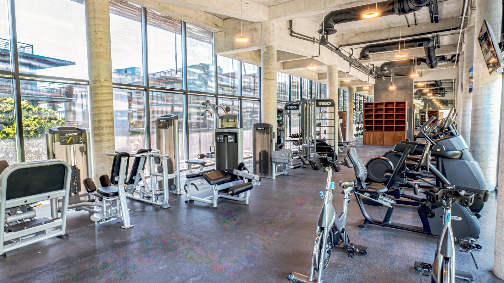 Ein Fitnessstudio mit unterschiedlichen Trainingsgeräten, großen Fenstern und einem Betonboden.