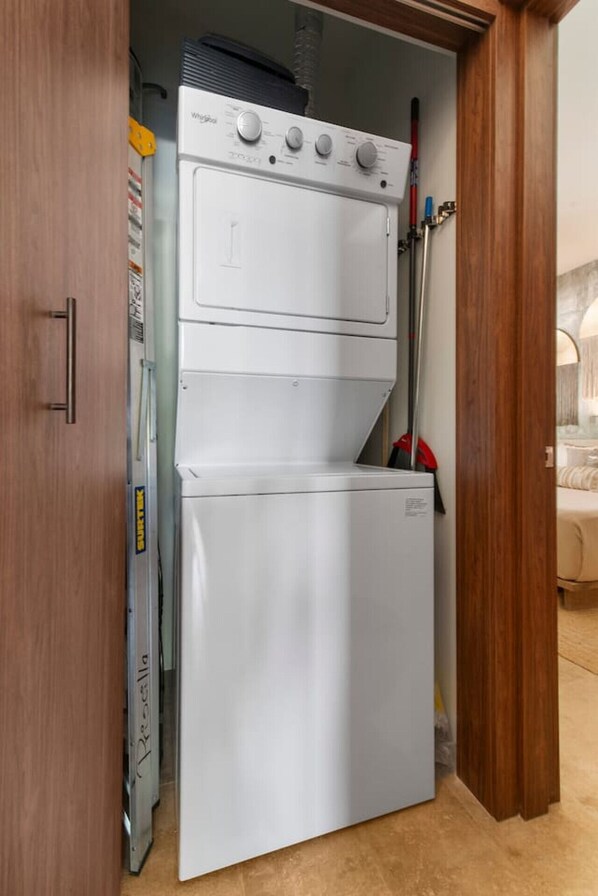 A compact laundry room with a white washing machine and dryer, a broom, and a vacuum cleaner. - Tulum 3BR Swim-Out Condo | 3 King Beds & Private Pool Access (Tulum)