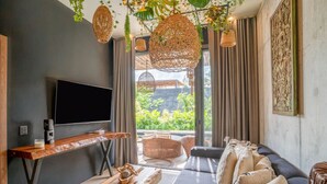 Un salón moderno con un televisor de pantalla plana grande, una mesa de centro de madera y un sofá con cojines. Hay lámparas colgantes de mimbre y vistas al exterior con vegetación.