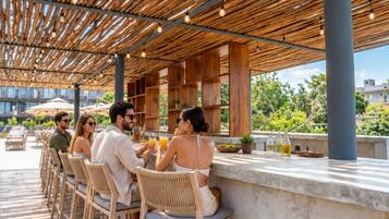 Ein Sitzbereich im Freien mit Holztischen und -stühlen, einer Pergola darüber und Blick auf viel Grün im Hintergrund.
