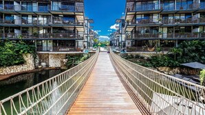 Eine Holzbrücke verbindet zwei moderne Gebäude mit Balkonen, umgeben von Grün und blauem Himmel.