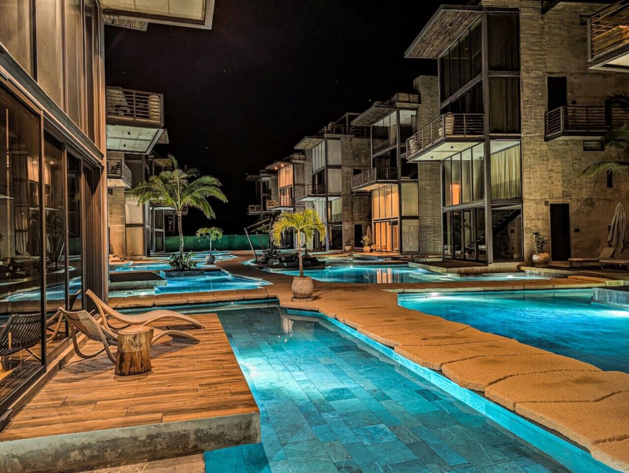 Nattebillede af et hotel med flere swimmingpools, træterrasser og moderne arkitektur.
