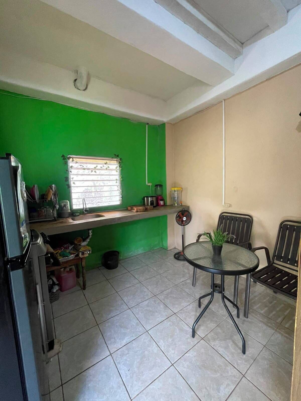 A compact kitchen with a green wall, a window with blinds, a sink, a refrigerator, a small dining table with chairs, and a fan.