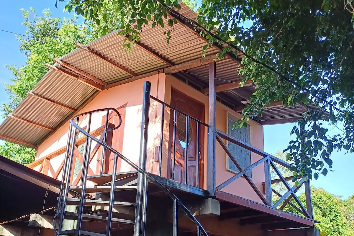 Una casa a due piani con un balcone, una scala e un'auto rossa parcheggiata davanti.