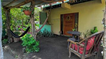 Un espacio al aire libre techado con una hamaca, una silla de madera con cojín rojo y una mesita.