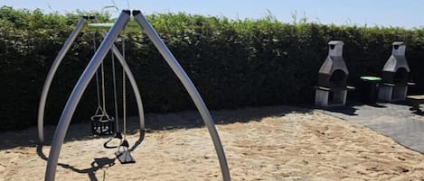 A swing set in a sandy outdoor area with a hedge in the background.