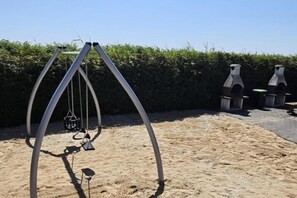 Spielplatz mit Schaukeln in einer sandigen Außenanlage, im Hintergrund ein Hecke.