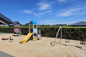 A playground with a slide and swings, surrounded by a hedge and a building with a sloped roof. - Apartment With a Balcony, Garden With Barbecue (Krynica Morska)