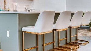 Een bar met een lichtblauwe toonbank, witte barkrukken met gouden poten, en een moderne keuken op de achtergrond.