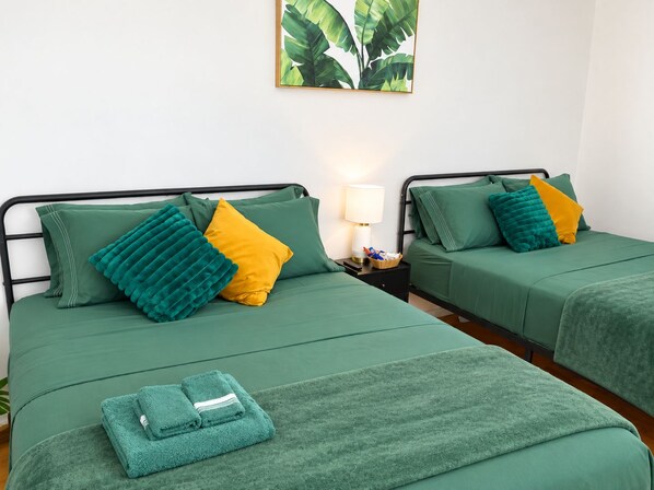 A hotel room with two beds, each with green bedding and multiple pillows, a bedside table with a lamp, and a framed plant on the wall.