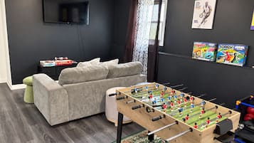 Olohuoneessa on pöytäjalkapallopeli, sohva, televisio ja ikkuna, jossa on verhot.