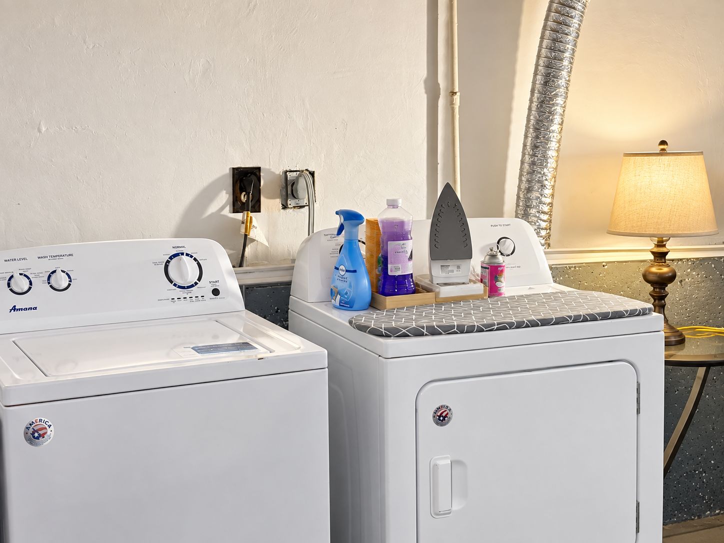 A laundry area with two front-loading washing machines, a mirror, a lamp, and various cleaning supplies.