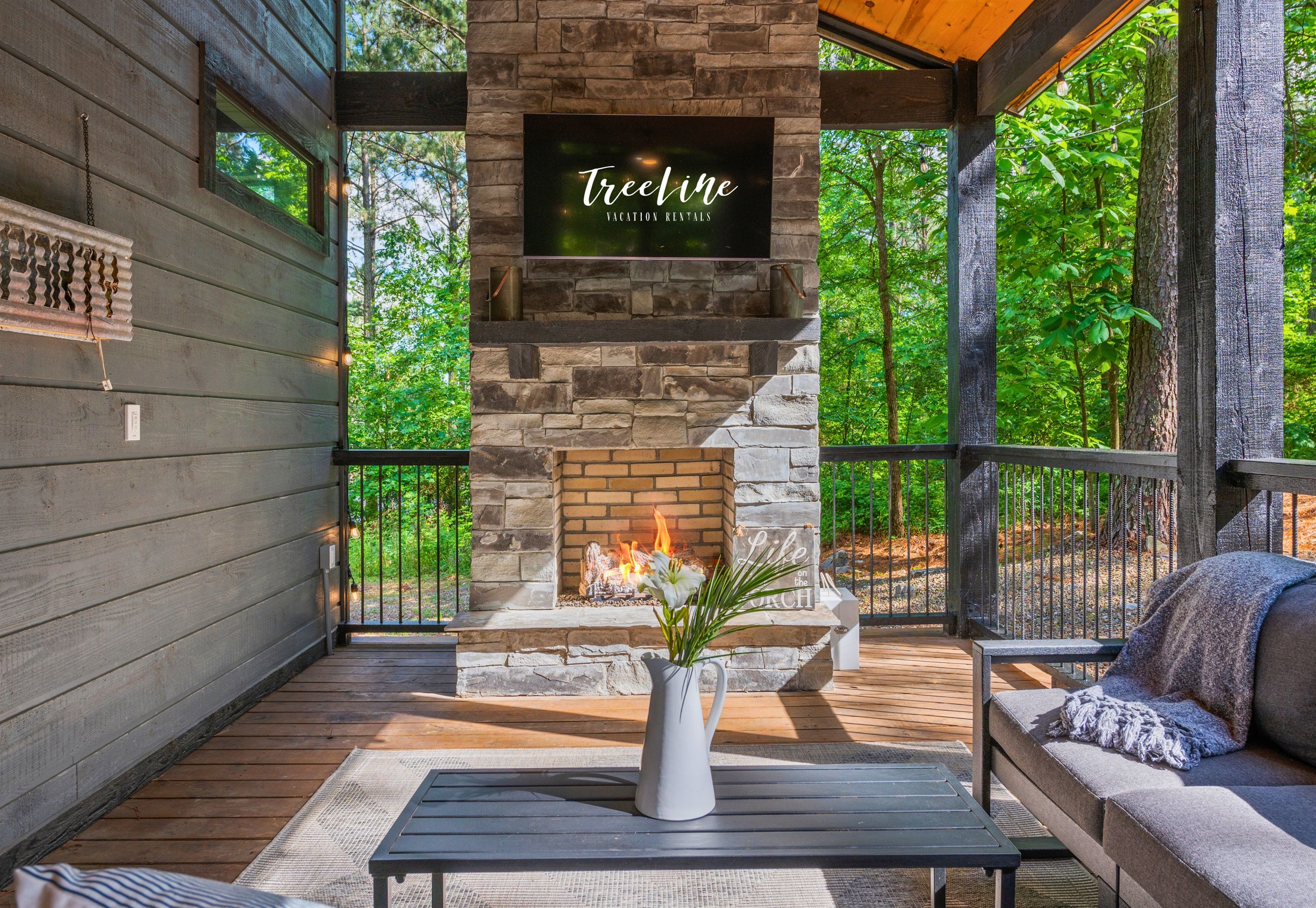 A cozy outdoor seating area with a stone fireplace, a TV mounted above it, and a wooden deck.