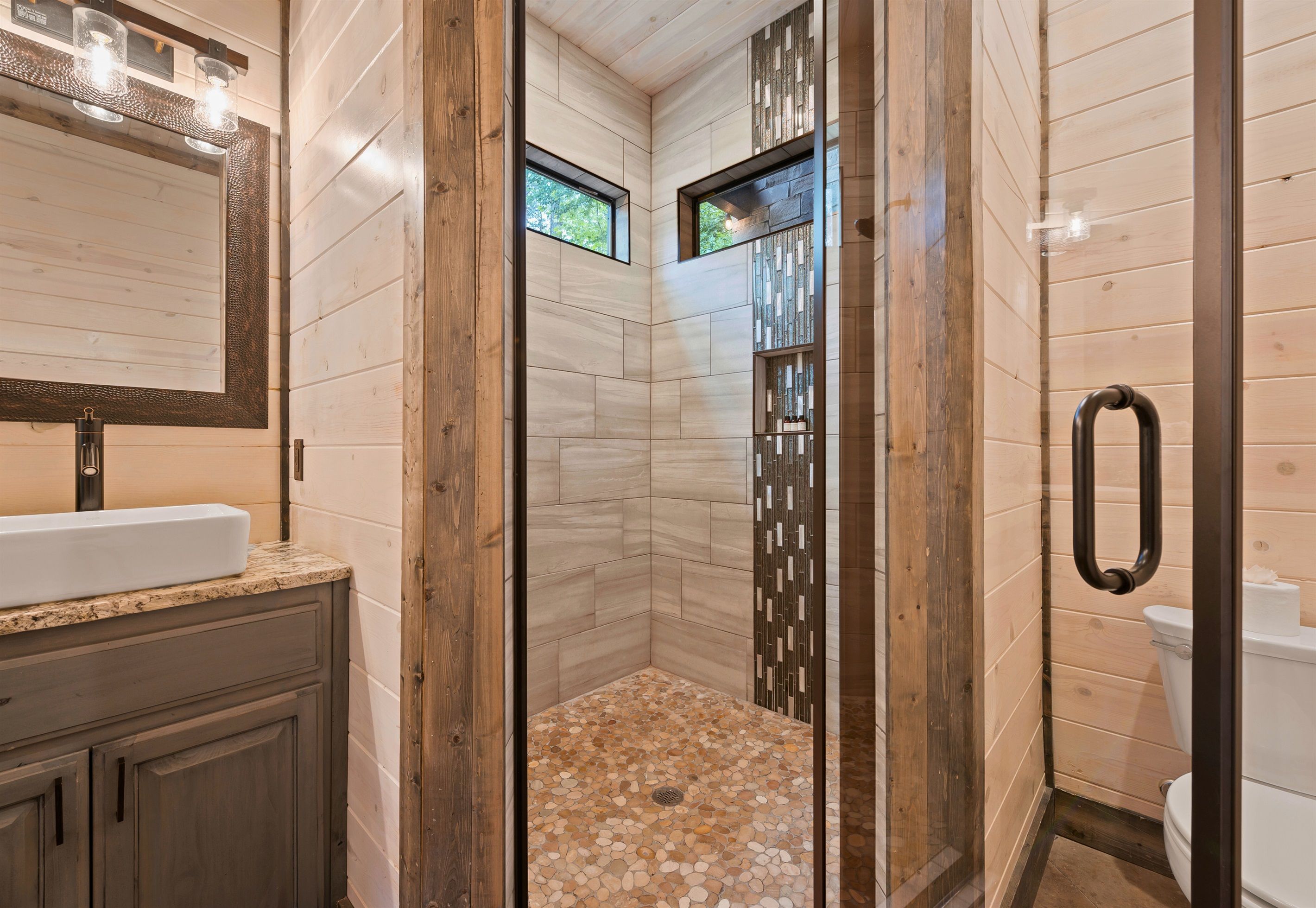 A bathroom with a wooden vanity, a large mirror, a shower with a pebble floor, and a toilet.