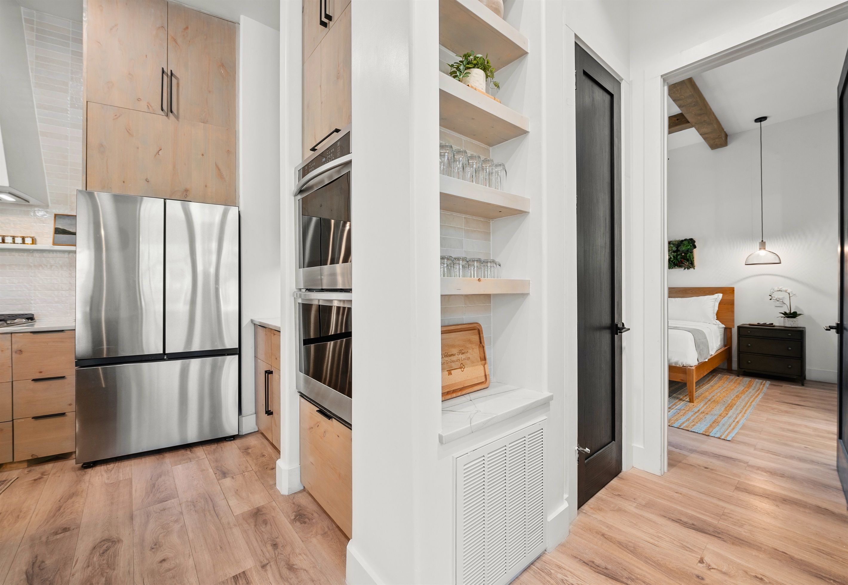 A modern kitchen with a stainless steel refrigerator, oven, and wooden cabinets. There is a white shelving unit with glassware and a plant. The kitchen leads to a bedroom with a bed and a nightstand.