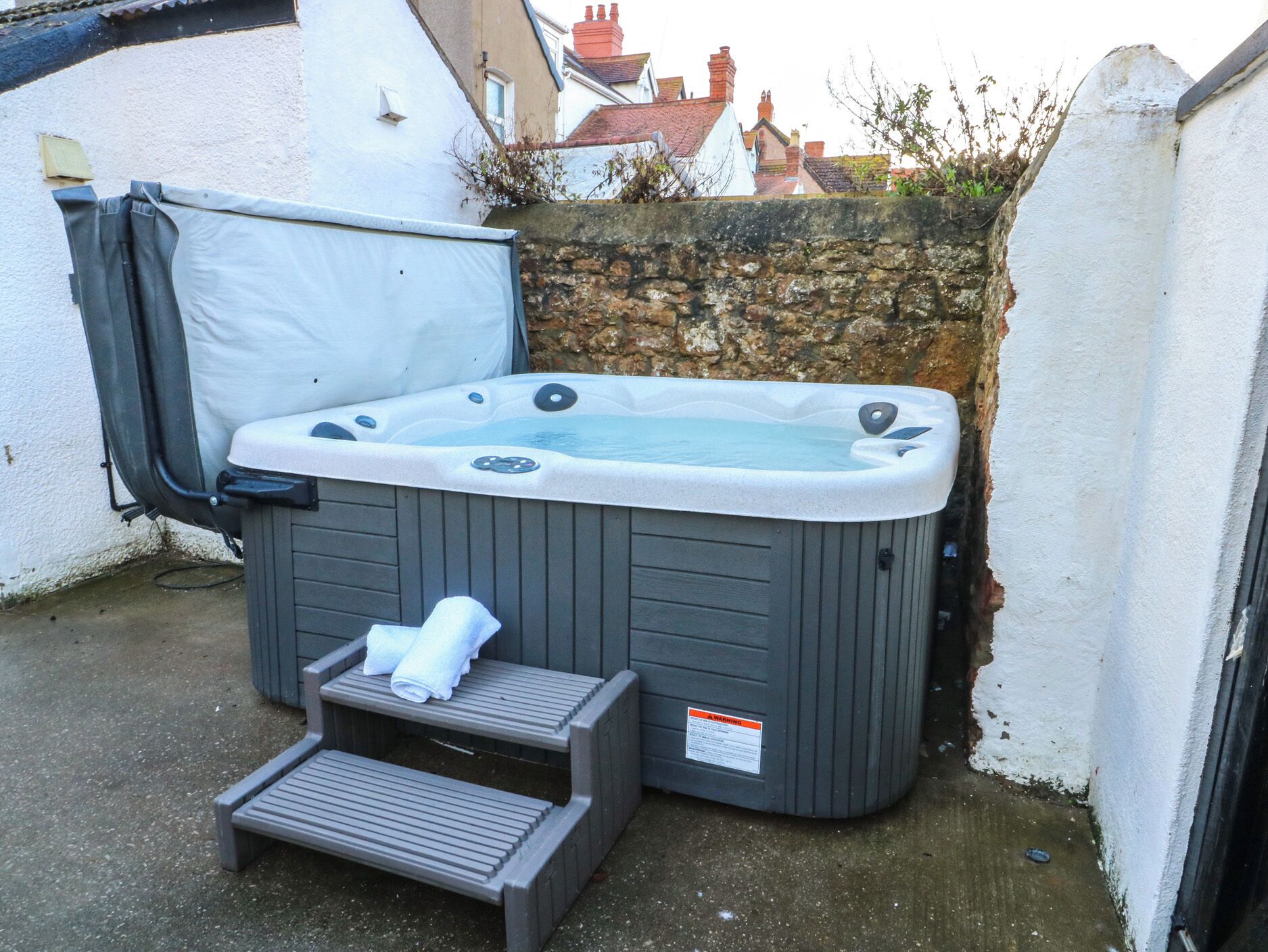 A hot tub with a step and a towel, set against a stone wall and a white building.