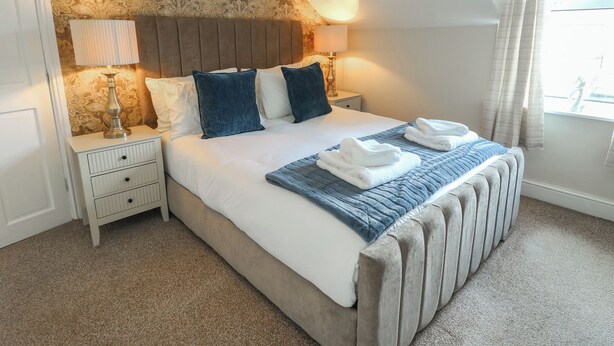 A neatly arranged bedroom with a large bed, two bedside tables, a lamp, and a view of the outdoors. - Curzon Heights (Llandudno)