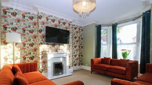 A living room with a fireplace, orange sofas, a flat-screen TV, floral wallpaper, and a large window with curtains. - Curzon Heights (Llandudno)