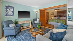 Una sala de estar con sofá, sillón y mesa de centro, un televisor montado en la pared, y una cama con cabecera de madera al fondo.