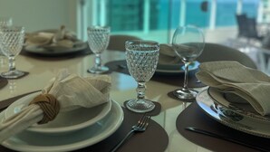 A dining table set with plates, glasses, and cutlery, with a view of a cityscape through a window. - Premium Ocean View Penthouse in Meia Praia - 12th floor, high standard, one block from the beach. (Itapema)
