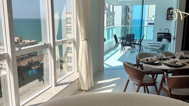 A modern living room with large windows, a dining area, and a view of the sea. - Premium Ocean View Penthouse in Meia Praia - 12th floor, high standard, one block from the beach. (Itapema)
