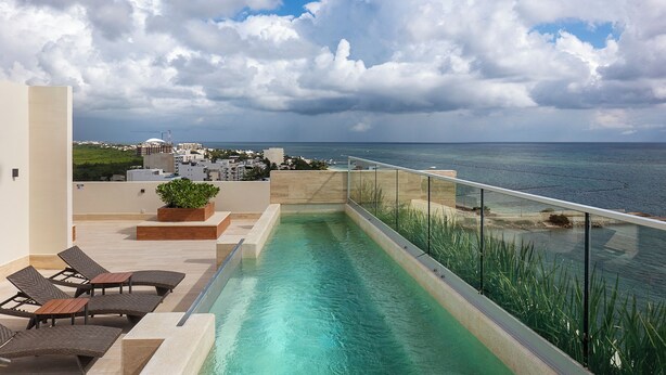 Ein Pool auf dem Dach mit Glasgeländer, Liegestühlen und Blick auf das Meer und die Stadt.