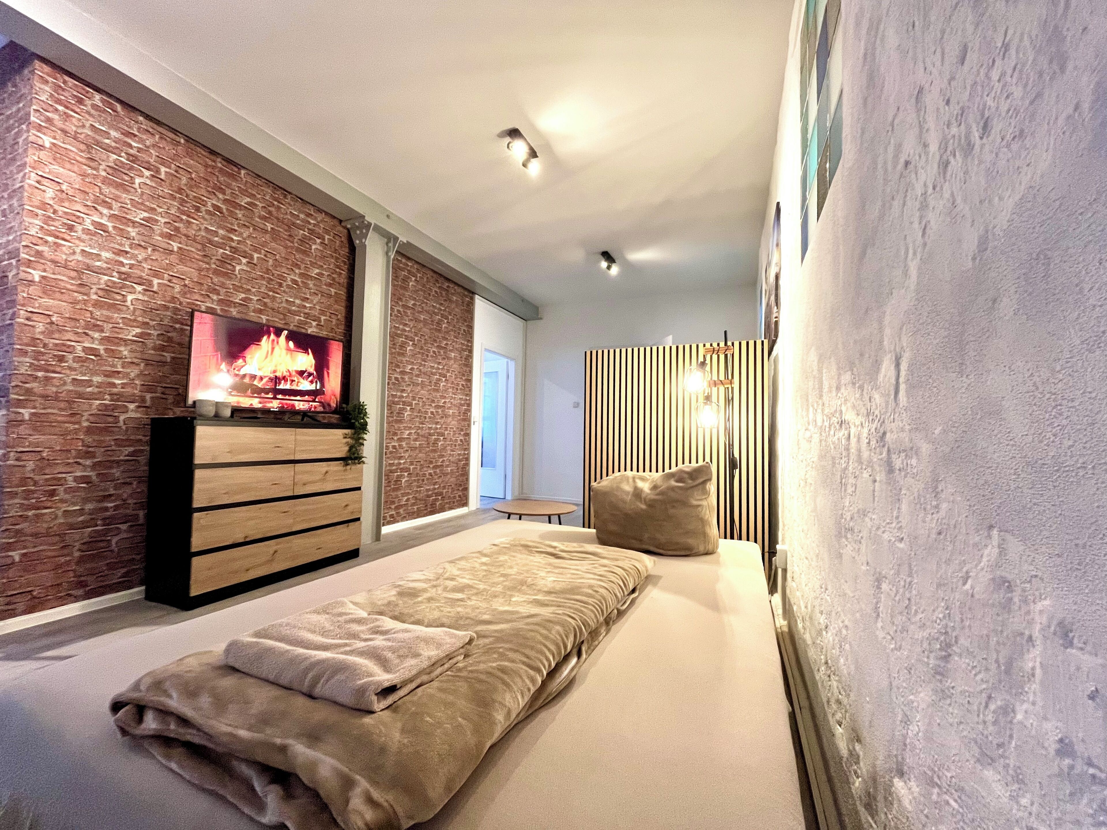 A modern bedroom with a brick wall, a TV, a bed with a beige blanket, and a wooden dresser.