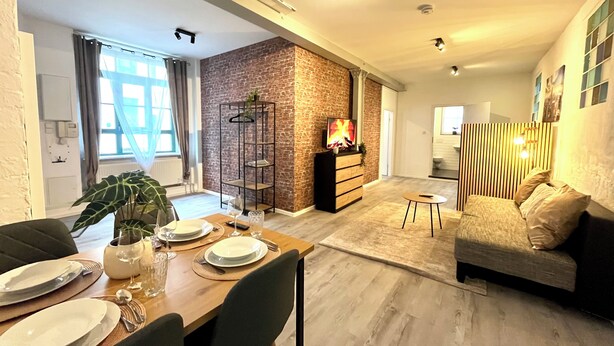Un salon moderne avec un coin repas, comprenant un mur d’accent en briques, une table à manger en bois pour quatre personnes, un canapé et un téléviseur posé sur un meuble en bois.