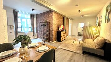 Una sala de estar moderna con comedor, que presenta una pared de ladrillo decorativa, una mesa de madera dispuesta para cuatro personas, un sofá y un televisor sobre un mueble de madera.