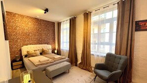 Une chambre avec un mur en briques, un lit doté d’une tête de lit capitonnée, un fauteuil gris et une table de chevet avec une bougie.