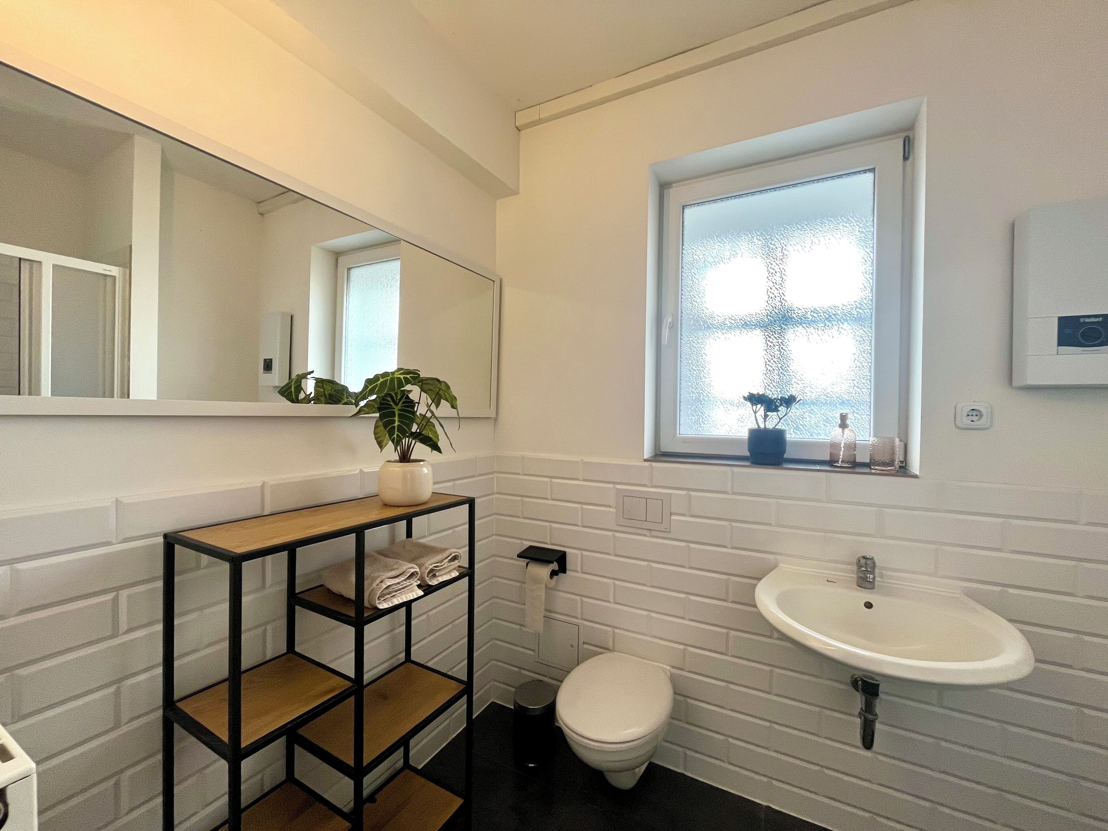 Un baño moderno con pared de azulejos blancos, tocador de madera con espejo, lavabo blanco y ventana con vistas.