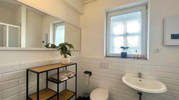 Un baño moderno con pared de azulejos blancos, tocador de madera con espejo, lavabo blanco y ventana con vistas.
