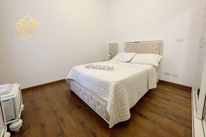 Ein ordentlich bezogenes Bett mit Decke, ein Nachttisch und ein Nachtschränkchen in einem Zimmer mit Holzboden und weißen Wänden.
