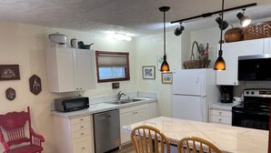 Cocina compacta con armarios blancos, lavavajillas, microondas y estufa. Hay dos sillas de madera junto a una mesa pequeña.