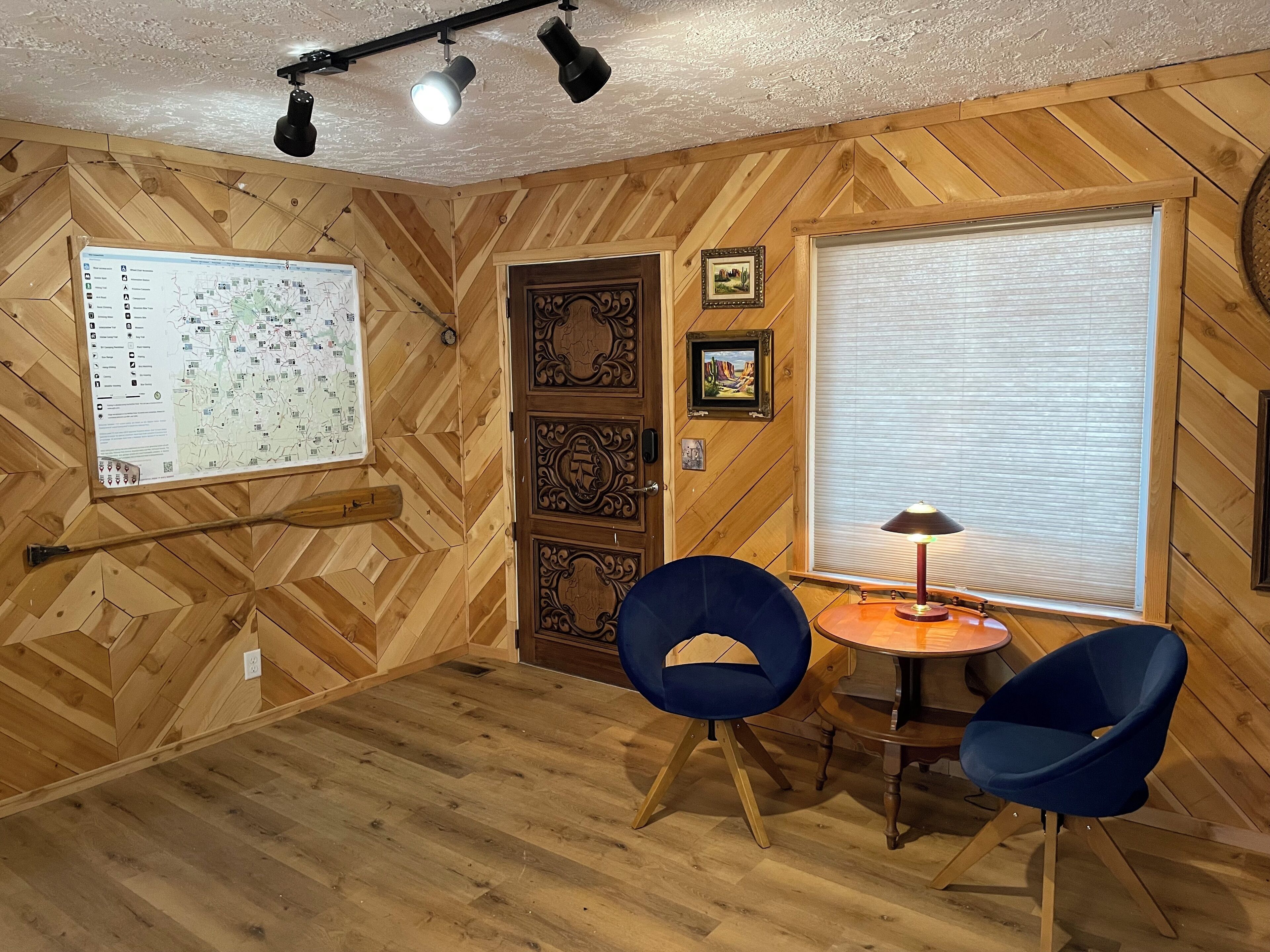 Una habitación con paredes de madera y un mapa en la pared, una puerta, una ventana con persianas y dos sillas azules alrededor de una pequeña mesa.