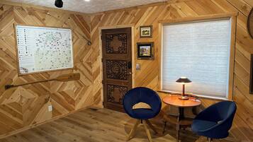 Una habitación con paredes de madera y un mapa en la pared, una puerta, una ventana con persianas y dos sillas azules alrededor de una pequeña mesa.