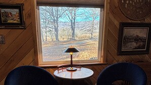 Un cuarto acogedor con paredes de madera, dos sillas azules, una mesita redonda con una lámpara y un ventanal con vistas a árboles y un cielo despejado.