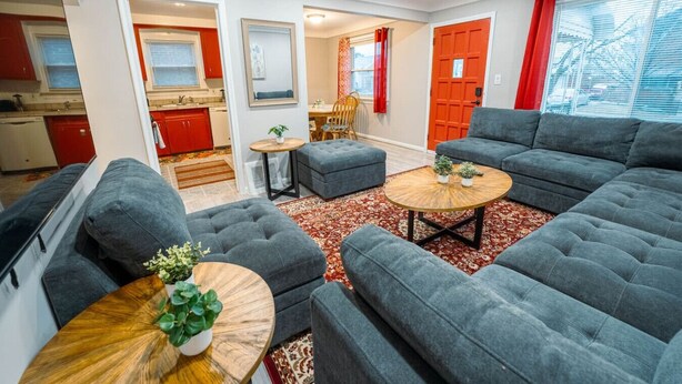 Ein modernes Wohnzimmer mit grauen Sofas, einem runden Couchtisch und einem roten, gemusterten Teppich. Im Hintergrund ist eine Küche mit roten Schränken und einem Fenster erkennbar.