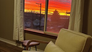 A room with a view of a sunset through a window, a beige armchair, and wooden flooring. - 2-bedroom studio sunny bright north hill location. (Pendleton)