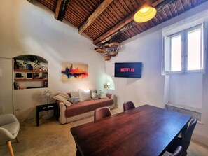 A cozy living room with a wooden ceiling, a sofa, a dining table, and a TV mounted on the wall. - Casa Bella in Central Noto, a Unique Sicilian Stay (Noto)