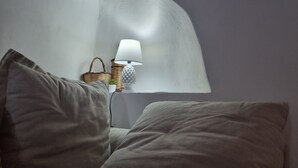 Un dormitorio con cama, almohadas, una lámpara, una planta pequeña y una cesta de mimbre.